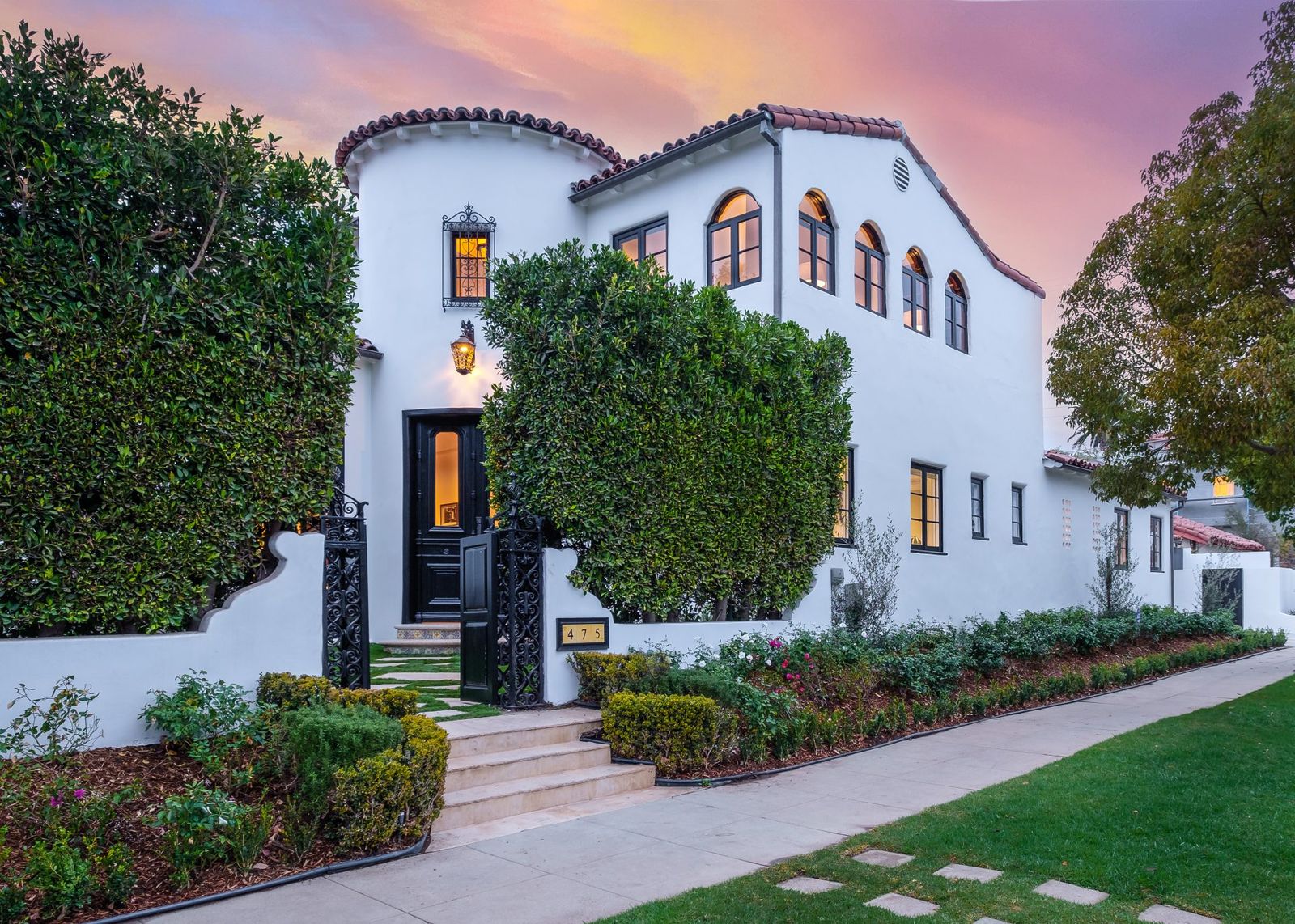 White Spanish revival house with a tile roof, arched windows and a landscaped front at dusk
