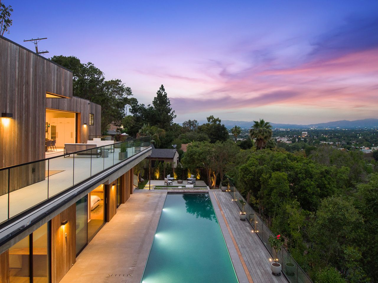 Modern hillside home with wood cladding, a glass-railed terrace and a long lit pool at sunset