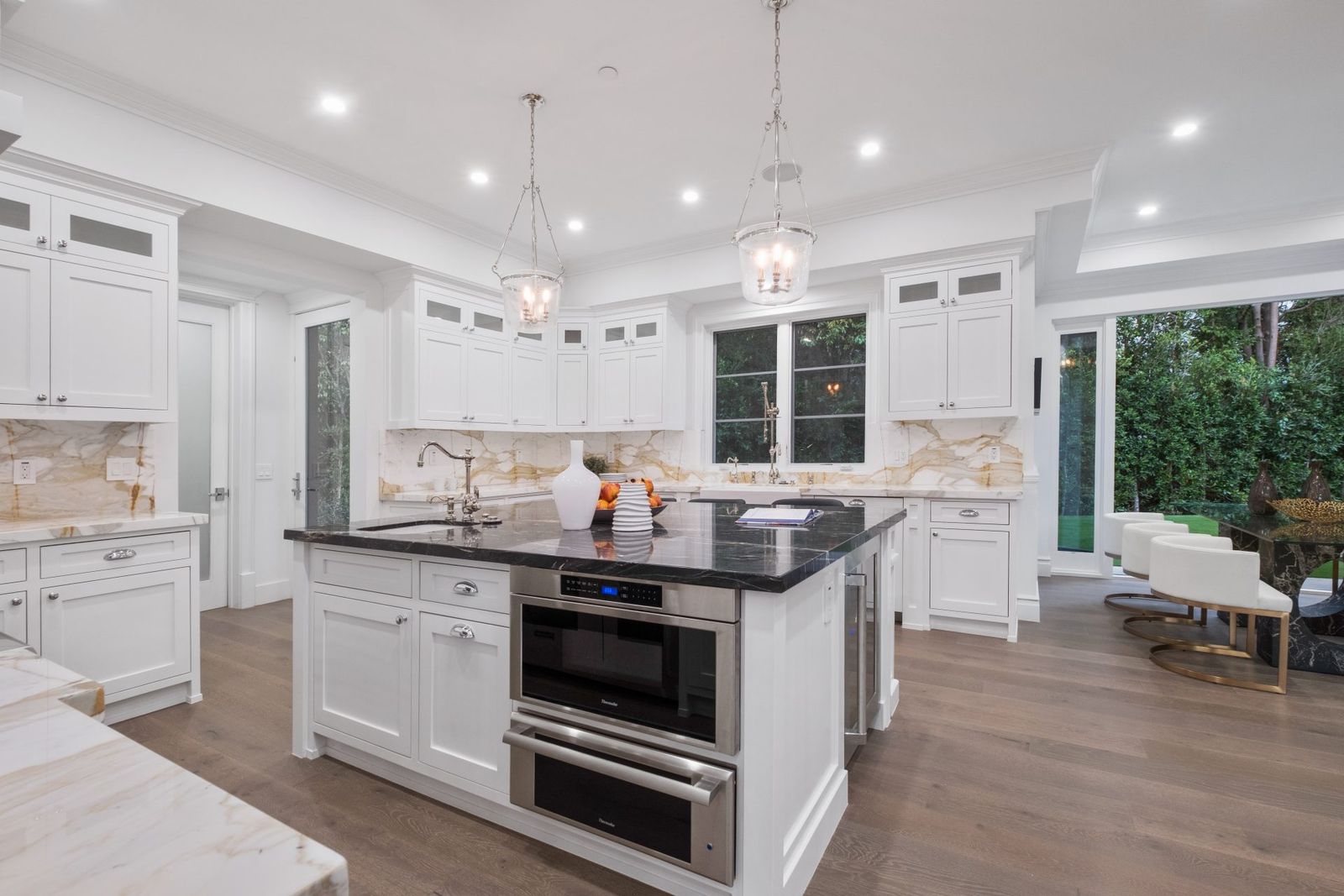 White transitional kitchen with a dark stone island, two glass pendant lights and windows onto a garden