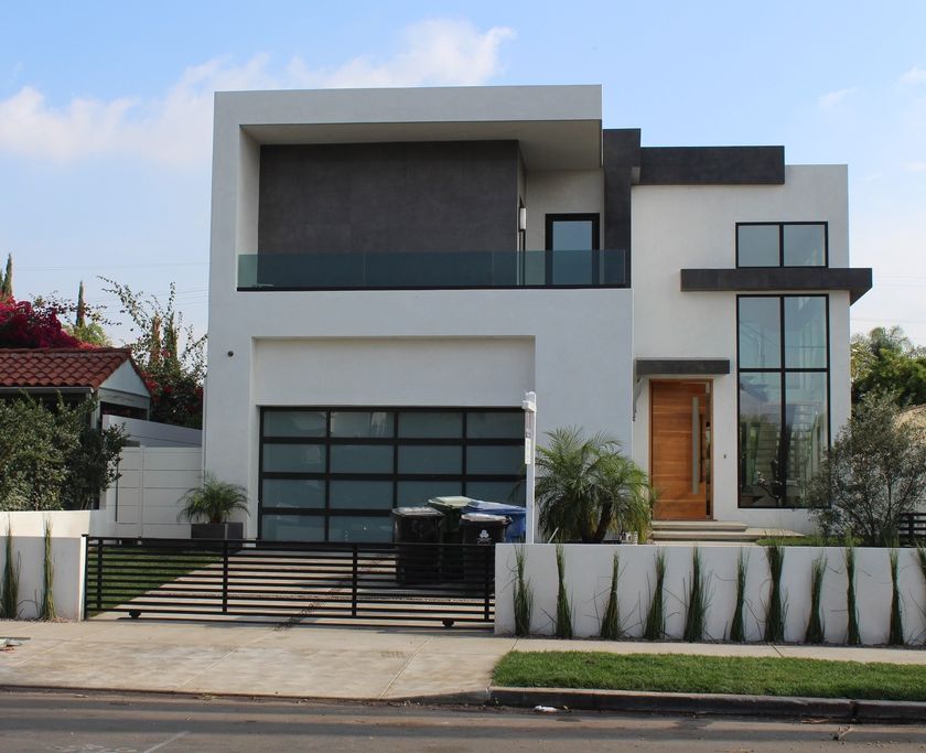 Two-story modern white house with a dark stone accent wall, a glass balcony rail, a black-framed glass garage door and a wood front door